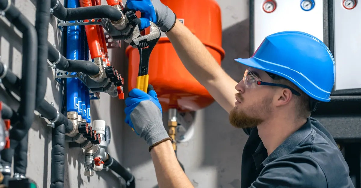 A plumber in a hard hat, safety glasses and gloves tightening a pipe fitting with an adjustable wrench beside a red expansion tank